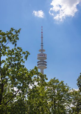 Münih, Almanya - 6 Temmuz 2022: Olympia Parkı 'ndaki Olympia Kulesi. Parkın ağaçlarından yukarı bakıyorum. Mavi gökyüzü ve yüksek kulenin arkasındaki beyaz bulut. Olimpiyat oyunları için yapılmış.