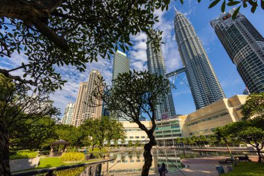 Kuala Lumpur, Malaysia - August 13, 2022: View of the Twin Towers through the palm trees of KLCC Park. Urban oasis with a fountain show. Petronas Towers the tallest twin skyscrapers in the world.