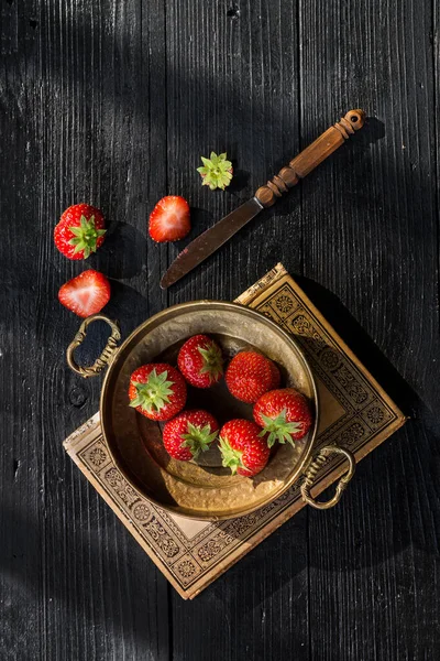 Fresh strawberries in vintage bronze bowl on a vintage old book on dark wooden background taken with side lighting
