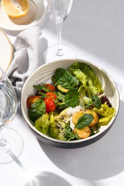 Avokado, domates ve peynir toplu salata kasesi. Sağlıklı vejetaryen yemeği ve beyaz masada su bardağı. Yaz yemeği, güneş ışığı ve sert gölge. Sağlıklı salata kasesi yemeği. Organik salata kasesi