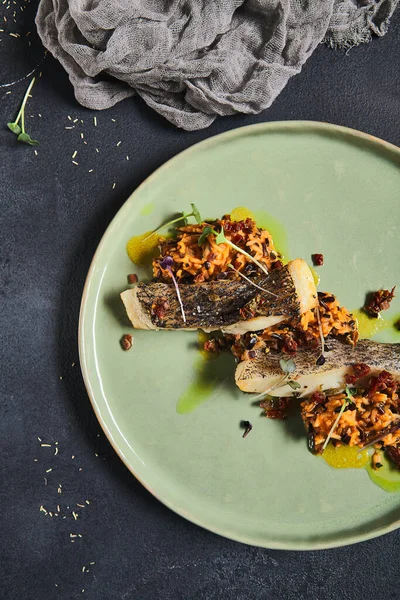 Risotto with slice roasted white fish in green plate on black background. Grilled cod with mix brown and wild rice in dark style. Minimal composition: fish and risotto on black table with textile