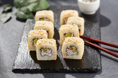 Karanlık taş masada Maki suşisi. Suşi rulo yengeç, peynir, salatalık içeride ve tamago dışarıda. Siyah arka planı, yaprakları ve sert gölgesi olan Japon tarzı bir menü.
