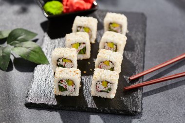 Karanlık taş masada Maki suşisi. Sushi susamlı California 'da yuvarlanır. Yengeçli Maki suşi, avokado, içinde salatalık, dışında susam. Siyah arka planı, yaprakları ve sert gölgesi olan Japon tarzı bir menü.