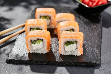 Karanlık taş masada Maki suşisi. Somonlu Philadelphia maki. Peynirli suşi, içinde salatalık, dışında somon. Siyah arka planı, yaprakları ve sert gölgesi olan Japon tarzı bir menü.