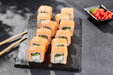 Karanlık taş masada Maki suşisi. Somonlu Philadelphia maki. Peynirli suşi, içinde salatalık, dışında somon. Siyah arka planı, yaprakları ve sert gölgesi olan Japon tarzı bir menü.
