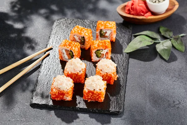 Maki sushi on dark stone table. California maki with tobiko. Sushi roll with salmon, cucumber inside, maguro, crab outside. Style concept japanese menu with black background, leaves and hard shadow