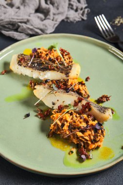 Siyah arka planda yeşil tabakta dilimlenmiş beyaz balıklı risotto. Kahverengi ve yabani pirinç karışımlı ızgara morina balığı. Asgari bileşim: Tekstil ile siyah masada balık ve risotto
