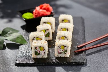 Karanlık taş masada Maki suşisi. Sushi susamlı California 'da yuvarlanır. Yengeçli Maki suşi, avokado, içinde salatalık, dışında susam. Siyah arka planı, yaprakları ve sert gölgesi olan Japon tarzı bir menü.