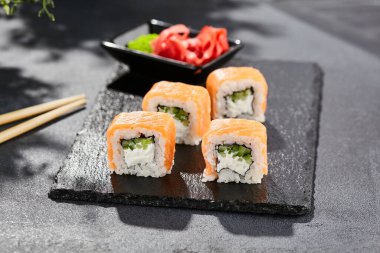 Karanlık taş masada Maki suşisi. Somonlu Philadelphia maki. Peynirli suşi, içinde salatalık, dışında somon. Siyah arka planı, yaprakları ve sert gölgesi olan Japon tarzı bir menü.