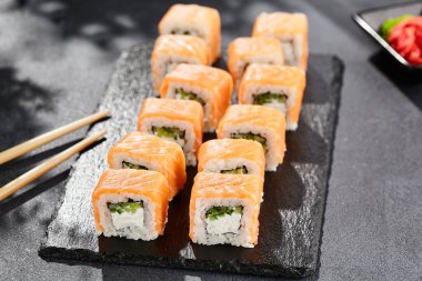 Karanlık taş masada Maki suşisi. Somonlu Philadelphia maki. Peynirli suşi, içinde salatalık, dışında somon. Siyah arka planı, yaprakları ve sert gölgesi olan Japon tarzı bir menü.