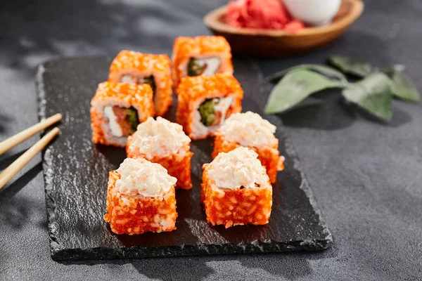 Maki sushi on dark stone table. California maki with tobiko. Sushi roll with salmon, cucumber inside, maguro, crab outside. Style concept japanese menu with black background, leaves and hard shadow