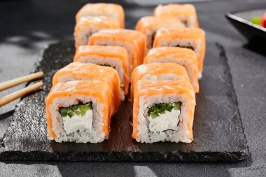 Karanlık taş masada Maki suşisi. Somonlu Philadelphia maki. Peynirli suşi, içinde salatalık, dışında somon. Siyah arka planı, yaprakları ve sert gölgesi olan Japon tarzı bir menü.