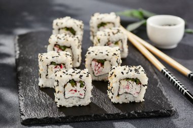 Kara tahtada Maki suşisi. Susamlı California maki. Yengeçli suşi, içinde salatalık, dışında susam. Siyah arka planı, yaprakları ve sert gölgesi olan Japon tarzı bir menü.