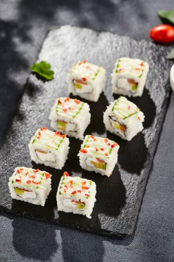 Koyu taş masada taze maki suşi. Sebzeli renkli rulo. Somonlu suşi, içinde avokado, dışında salatalık. Siyah arka planı, yaprakları ve sert gölgesi olan Japon tarzı bir menü.