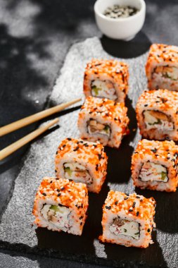 Kara tahtada Maki suşisi. Masaj ve susamlı California maki 'si. Yengeçli suşi, içeride salatalık, dışarıda tobiko. Siyah arka planı, yaprakları ve sert gölgesi olan Japon tarzı bir menü.