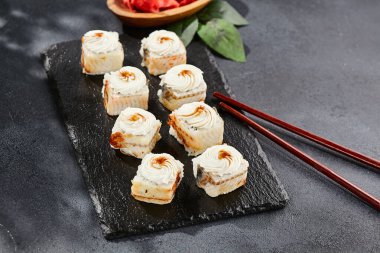 Kara tahtada Maki suşisi. Yılanbalıklı Unagi maki. Yılanbalığı, krem peynir ve unagi soslu suşi. Siyah arka planı, yaprakları ve sert gölgesi olan Japon tarzı bir menü.