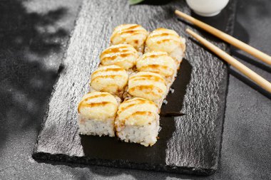 Kara tahtada fırında Maki suşisi. Unagi soslu sıcak peynirli maki. Üstünde fırında peynir ve unagi soslu suşi rulosu. Siyah arka planı, yaprakları ve sert gölgesi olan Japon tarzı bir menü.