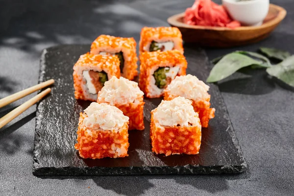 Maki sushi on dark stone table. California maki with tobiko. Sushi roll with salmon, cucumber inside, maguro, crab outside. Style concept japanese menu with black background, leaves and hard shadow