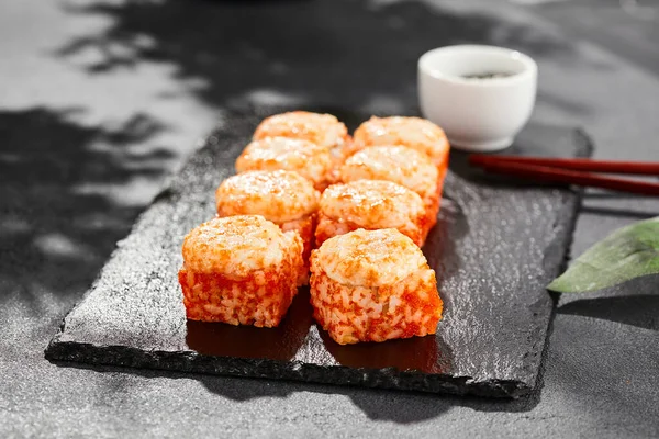 Baked Maki sushi on dark slate. Hot california maki with tobiko. Sushi roll with masago caviar, cheese topped baked oven. Style concept japanese menu with black background, leaves and hard shadow