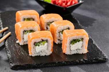 Karanlık taş masada Maki suşisi. Somonlu Philadelphia maki. Peynirli suşi, içinde salatalık, dışında somon. Siyah arka planı, yaprakları ve sert gölgesi olan Japon tarzı bir menü.