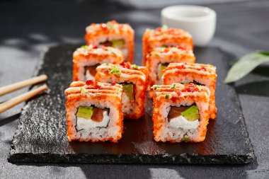 Karanlık taş masada Maki suşisi. Tobiko ile Kaliforniya maki 'si. Peynirli suşi, somon, içinde avokado, dışında maguro. Siyah arka planı, yaprakları ve sert gölgesi olan Japon tarzı bir menü.
