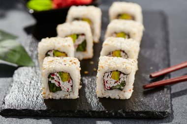 Karanlık taş masada Maki suşisi. Sushi susamlı California 'da yuvarlanır. Yengeçli Maki suşi, avokado, içinde salatalık, dışında susam. Siyah arka planı, yaprakları ve sert gölgesi olan Japon tarzı bir menü.