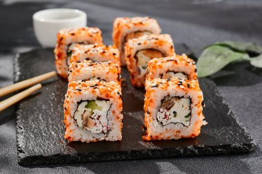 Kara tahtada Maki suşisi. Masaj ve susamlı California maki 'si. Yengeçli suşi, içeride salatalık, dışarıda tobiko. Siyah arka planı, yaprakları ve sert gölgesi olan Japon tarzı bir menü.