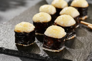 Kara tahtada fırında Maki suşisi. Sıcak peynirli hosomaki rulosu. Peynirli, fırında peynirli sade suşi. Siyah arka planı, yaprakları ve sert gölgesi olan Japon tarzı bir menü.