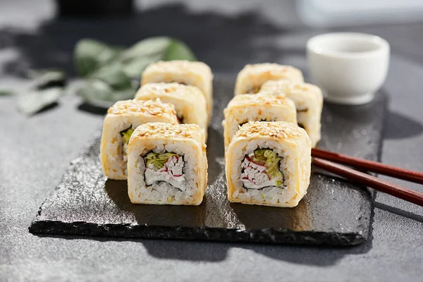 Karanlık taş masada Maki suşisi. Suşi rulo yengeç, peynir, salatalık içeride ve tamago dışarıda. Siyah arka planı, yaprakları ve sert gölgesi olan Japon tarzı bir menü.