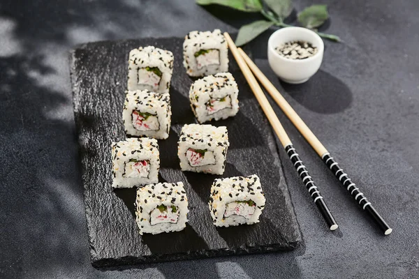 Kara tahtada Maki suşisi. Susamlı California maki. Yengeçli suşi, içinde salatalık, dışında susam. Siyah arka planı, yaprakları ve sert gölgesi olan Japon tarzı bir menü.