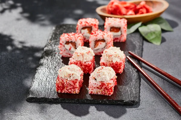 Kara tahtada Maki suşisi. Yengeçli Maki dürümü. Yengeç, krem peynir ve tobika ile suşi rulosu. Siyah arka planı, yaprakları ve sert gölgesi olan Japon tarzı bir menü.