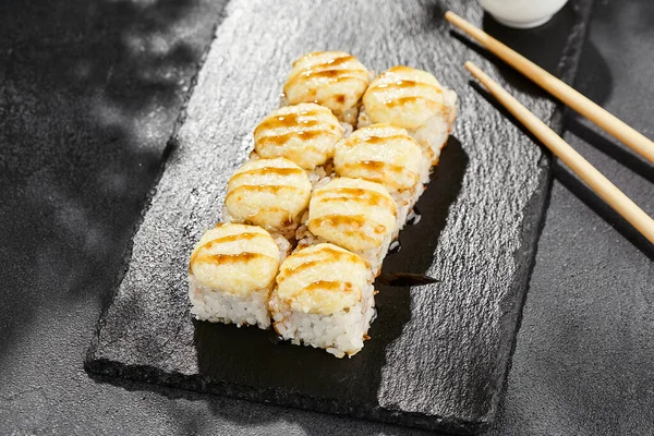 Kara tahtada fırında Maki suşisi. Unagi soslu sıcak peynirli maki. Üstünde fırında peynir ve unagi soslu suşi rulosu. Siyah arka planı, yaprakları ve sert gölgesi olan Japon tarzı bir menü.