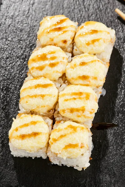 Kara tahtada fırında Maki suşisi. Unagi soslu sıcak peynirli maki. Üstünde fırında peynir ve unagi soslu suşi rulosu. Siyah arka planı, yaprakları ve sert gölgesi olan Japon tarzı bir menü.