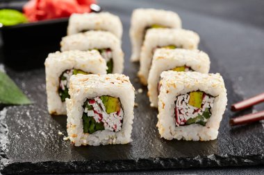 Karanlık taş masada Maki suşisi. Sushi susamlı California 'da yuvarlanır. Yengeçli Maki suşi, avokado, içinde salatalık, dışında susam. Siyah arka planı, yaprakları ve sert gölgesi olan Japon tarzı bir menü.