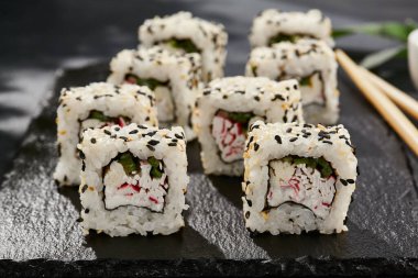 Kara tahtada Maki suşisi. Susamlı California maki. Yengeçli suşi, içinde salatalık, dışında susam. Siyah arka planı, yaprakları ve sert gölgesi olan Japon tarzı bir menü.