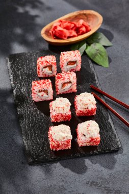 Kara tahtada Maki suşisi. Yengeçli Maki dürümü. Yengeç, krem peynir ve tobika ile suşi rulosu. Siyah arka planı, yaprakları ve sert gölgesi olan Japon tarzı bir menü.