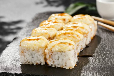 Kara tahtada fırında Maki suşisi. Unagi soslu sıcak peynirli maki. Üstünde fırında peynir ve unagi soslu suşi rulosu. Siyah arka planı, yaprakları ve sert gölgesi olan Japon tarzı bir menü.