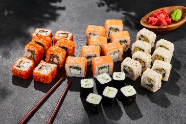 Maki suşisi karanlık tahtaya kurulmuş. Çeşitli ekmeklerle Maki seti. Suşi rulolarından - Philadelphia, California, hosomaki. Siyah arka planı, yaprakları ve sert gölgesi olan tarz konsept suşi menüsü