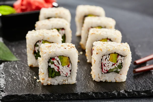 Karanlık taş masada Maki suşisi. Sushi susamlı California 'da yuvarlanır. Yengeçli Maki suşi, avokado, içinde salatalık, dışında susam. Siyah arka planı, yaprakları ve sert gölgesi olan Japon tarzı bir menü.