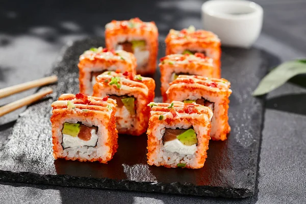 Karanlık taş masada Maki suşisi. Tobiko ile Kaliforniya maki 'si. Peynirli suşi, somon, içinde avokado, dışında maguro. Siyah arka planı, yaprakları ve sert gölgesi olan Japon tarzı bir menü.