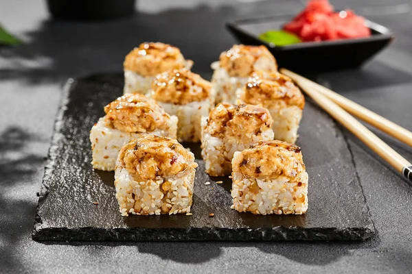 Baked Maki sushi on dark slate. Hot unagi maki with eel. Sushi roll with baked eel, cheese and unagi sauce topped. Style concept japanese menu with black background, leaves and hard shadow