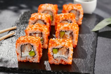 Karanlık taş masada Maki suşisi. Tobiko ile Kaliforniya maki 'si. Peynirli suşi, karides, içinde avokado, dışında maguro. Siyah arka planı, yaprakları ve sert gölgesi olan Japon tarzı bir menü.