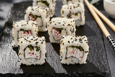 Kara tahtada Maki suşisi. Susamlı California maki. Yengeçli suşi, içinde salatalık, dışında susam. Siyah arka planı, yaprakları ve sert gölgesi olan Japon tarzı bir menü.