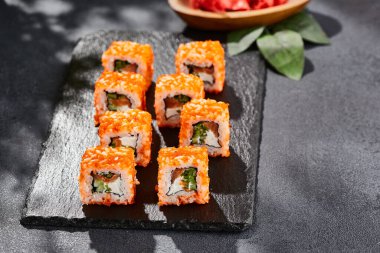 Kara tahtada Maki suşisi. Somonlu California maki 'si. Somonlu suşi, içinde salatalık, dışında tobiko. Siyah arka planı, yaprakları ve sert gölgesi olan Japon tarzı bir menü.