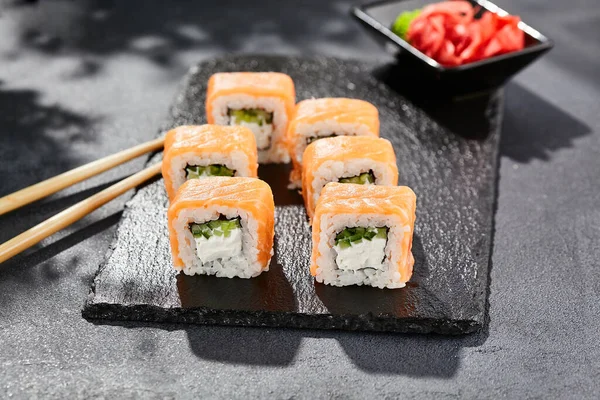 Karanlık taş masada Maki suşisi. Somonlu Philadelphia maki. Peynirli suşi, içinde salatalık, dışında somon. Siyah arka planı, yaprakları ve sert gölgesi olan Japon tarzı bir menü.