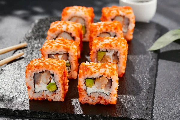 Maki sushi on dark stone table. California maki with tobiko. Sushi roll with cheese,shrimp, avocado inside, maguro outside. Style concept japanese menu with black background, leaves and hard shadow