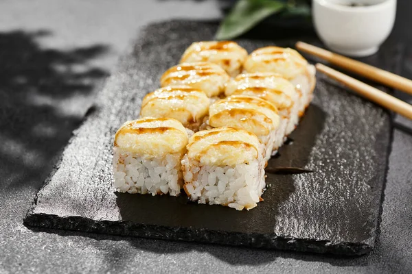 Kara tahtada fırında Maki suşisi. Unagi soslu sıcak peynirli maki. Üstünde fırında peynir ve unagi soslu suşi rulosu. Siyah arka planı, yaprakları ve sert gölgesi olan Japon tarzı bir menü.