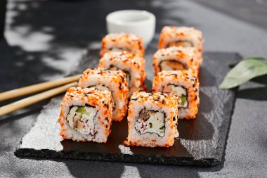 Kara tahtada Maki suşisi. Masaj ve susamlı California maki 'si. Yengeçli suşi, içeride salatalık, dışarıda tobiko. Siyah arka planı, yaprakları ve sert gölgesi olan Japon tarzı bir menü.