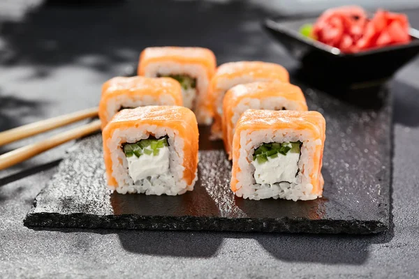 暗い石のテーブルの上に巻き寿司 フィラデルフィア巻きとサーモン 寿司ロールチーズ キュウリの内側 外側のサーモン 黒を基調としたスタイルのコンセプト日本料理 葉とハードシャドウ — ストック写真
