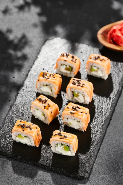 Kara tahtada Maki suşisi. Fırında somonlu sıcak Philadelphia maki. Izgara somon, krem peynir ve avokado ile suşi rulosu. Siyah arka planı, yaprakları ve sert gölgesi olan Japon tarzı bir menü.